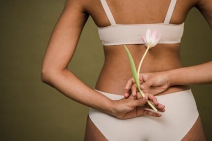 Back view of crop anonymous young female in white bra and panties holding fresh delicate tulip flower against beige background
